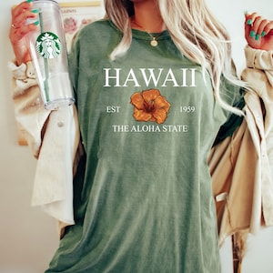 以下が含まれることがあります： ハワイ EST 1959 THE ALOHA STATE と書かれた緑色のTシャツと、黄色のハイビスカスの花柄のグラフィック