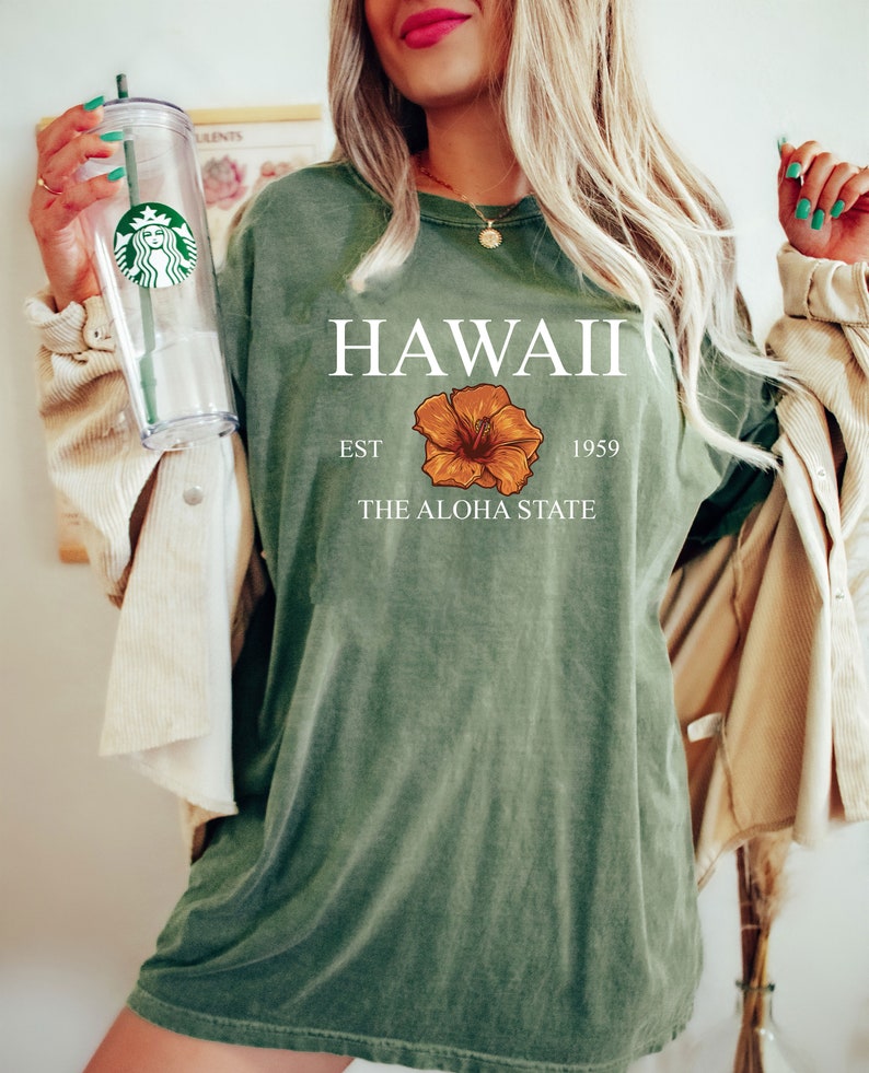 以下が含まれることがあります： ハワイ EST 1959 THE ALOHA STATE と書かれた緑色のTシャツと、黄色のハイビスカスの花柄のグラフィック