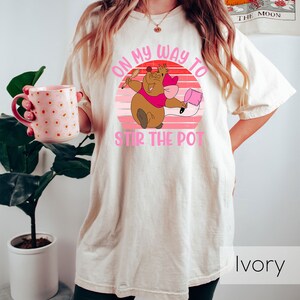 May include: A white t-shirt with a pink and brown graphic of a cartoon character holding a spoon and a pot. The graphic says "On my way to stir the pot". The shirt is ivory.