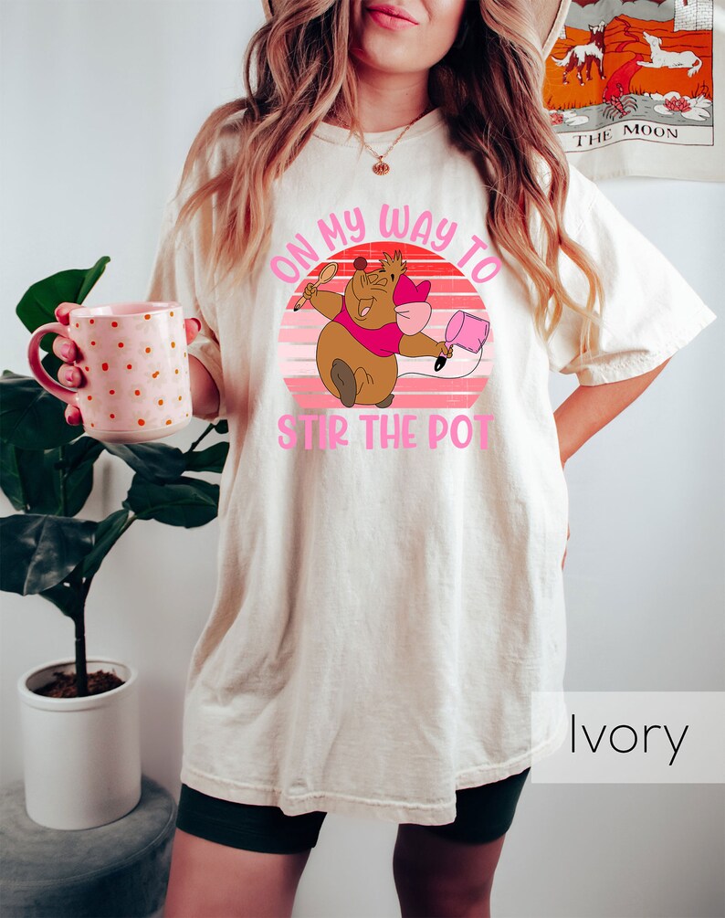 May include: A white t-shirt with a pink and brown graphic of a cartoon character holding a spoon and a pot. The graphic says "On my way to stir the pot". The shirt is ivory.