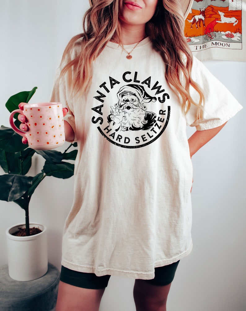 May include: Cream-colored t-shirt featuring a circular graphic with the text "SANTA CLAUS HARD SELTZER" and an illustration of Santa Claus. The shirt is a relaxed fit. A person is holding a pink mug with white polka dots.
