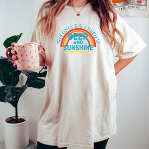 May include: Cream-colored t-shirt with the text "THE ONLY B.S. I NEED IS BEER AND SUNSHINE" in a rainbow design. The shirt is worn by a person holding a pink mug with orange polka dots. A plant is in the background.