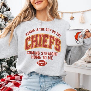 May include: A light gray sweatshirt with the text "Karma is the Guy on the Chiefs Coming Straight Home to Me" in red and gold. The sweatshirt features a football graphic. A white mug with a Santa Claus graphic is held in the hand.
