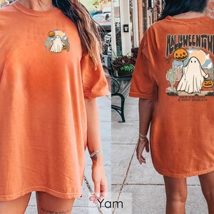 May include: Orange t-shirt with a Halloween-themed graphic. The front features a small design of a ghost and pumpkins. The back displays "Halloweentown" text with a ghost, pumpkins, and desert landscape. The shirt is a relaxed fit.