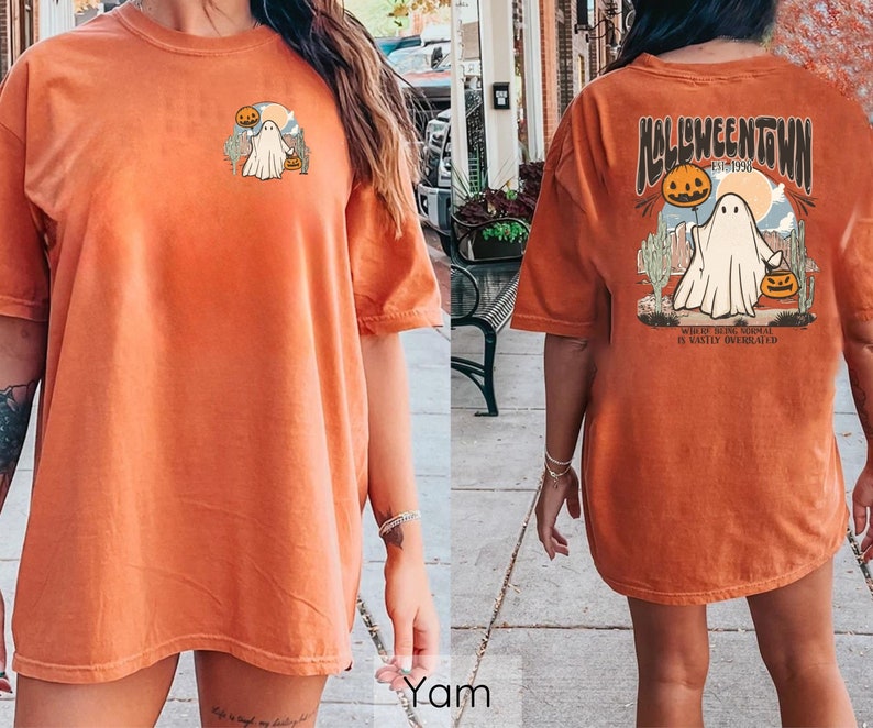 May include: Orange t-shirt with a Halloween-themed graphic. The front features a small design of a ghost and pumpkins. The back displays "Halloweentown" text with a ghost, pumpkins, and desert landscape. The shirt is a relaxed fit.