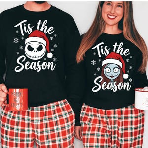 Pode incluir: Camisas pretas de manga comprida com o texto "Tis the Season" e desenhos de Jack Skellington e Sally, combinadas com cal&ccedil;as de pijama xadrez vermelhas. Uma caneca vermelha com texto &eacute; segurada.