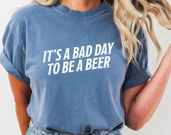Puede incluir: Una camiseta azul con la frase "IT'S A BAD DAY TO BE A BEER" en letras blancas. La camiseta es de color liso y tiene mangas cortas. La modelo lleva vaqueros azules.