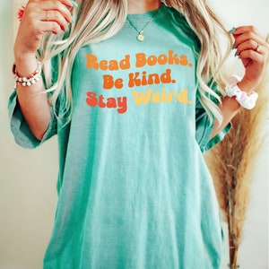 K&ouml;nnte beinhalten: &Uuml;bergr&ouml;&szlig;en-T-Shirt in T&uuml;rkisblau mit dem Text "Read Books. Be Kind. Stay Weird." in einer Retro-Schriftart mit orange, gelben und roten Farben.