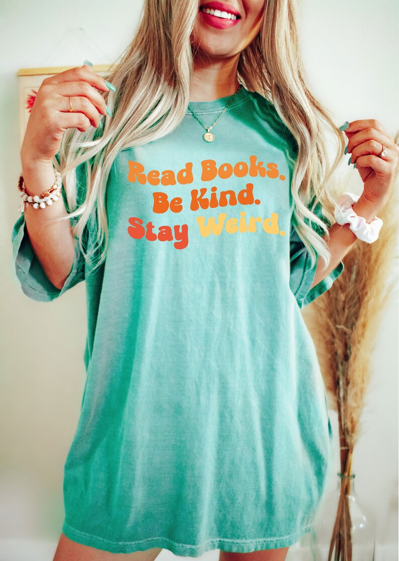 K&ouml;nnte beinhalten: &Uuml;bergr&ouml;&szlig;en-T-Shirt in T&uuml;rkisblau mit dem Text "Read Books. Be Kind. Stay Weird." in einer Retro-Schriftart mit orange, gelben und roten Farben.