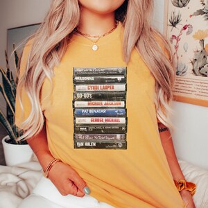 May include: Mustard yellow t-shirt with a print of stacked cassette tapes. The tapes show artist names including Madonna, Cyndi Lauper, and Michael Jackson. The t-shirt has a relaxed fit.