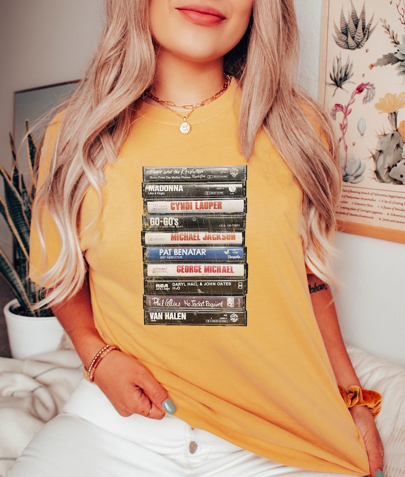 May include: Mustard yellow t-shirt with a print of stacked cassette tapes. The tapes show artist names including Madonna, Cyndi Lauper, and Michael Jackson. The t-shirt has a relaxed fit.
