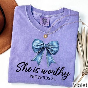 May include: A purple t-shirt with a blue denim bow and the text "She is worthy Proverbs 31" printed on the front.