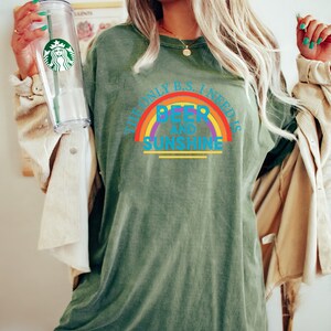 May include: Olive green t-shirt with the text "THE ONLY B.S. I NEED IS BEER AND SUNSHINE" in a rainbow design. The shirt is worn with a beige jacket and a clear Starbucks tumbler.