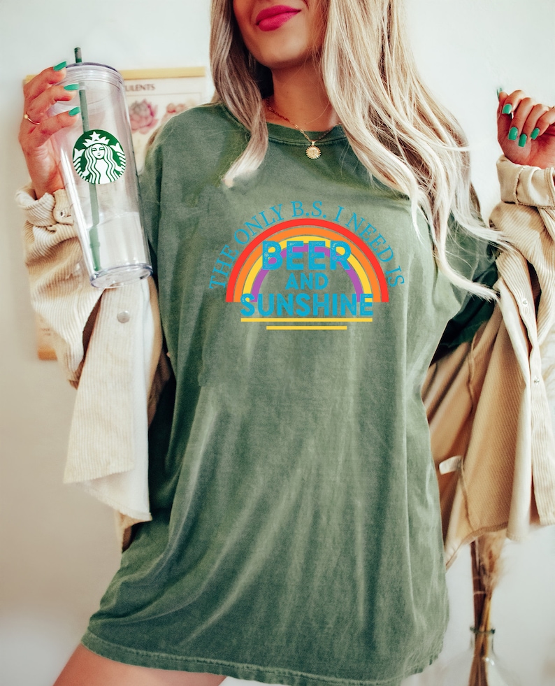 May include: Olive green t-shirt with the text "THE ONLY B.S. I NEED IS BEER AND SUNSHINE" in a rainbow design. The shirt is worn with a beige jacket and a clear Starbucks tumbler.