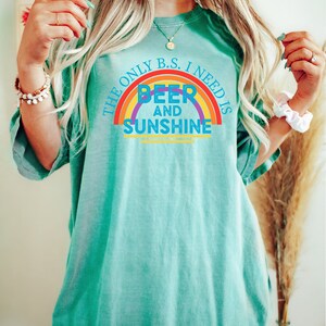 May include: A turquoise t-shirt with the text "THE ONLY B.S. I NEED IS BEER AND SUNSHINE" in a rainbow design. The shirt is a relaxed fit and has short sleeves.