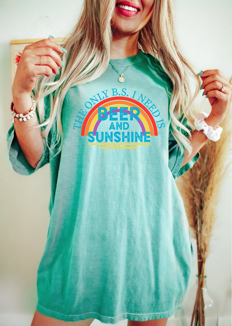 May include: A turquoise t-shirt with the text "THE ONLY B.S. I NEED IS BEER AND SUNSHINE" in a rainbow design. The shirt is a relaxed fit and has short sleeves.