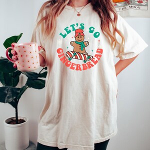 May include: Cream-colored t-shirt featuring a graphic of a gingerbread man on a skateboard, with the text "Let's Go Gingerbread" in green and red. The gingerbread man wears a red hat and scarf. The shirt is a relaxed fit.