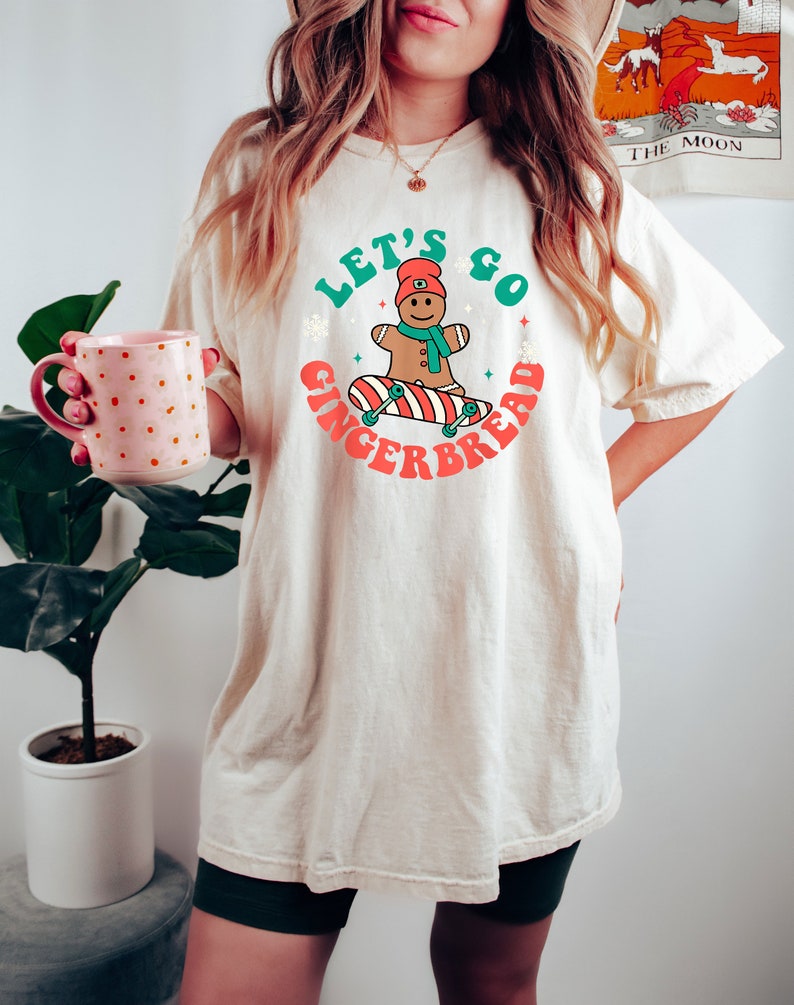 May include: Cream-colored t-shirt featuring a graphic of a gingerbread man on a skateboard, with the text "Let's Go Gingerbread" in green and red. The gingerbread man wears a red hat and scarf. The shirt is a relaxed fit.