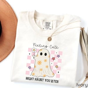 May include: Ivory-colored t-shirt with a ghost graphic holding a flower. The ghost is decorated with orange flowers and set against a pink and white checkered background. The text "Feeling Cute Might Haunt You Later" is printed on the shirt.