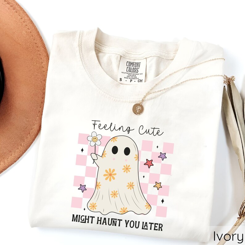 May include: Ivory-colored t-shirt with a ghost graphic holding a flower. The ghost is decorated with orange flowers and set against a pink and white checkered background. The text "Feeling Cute Might Haunt You Later" is printed on the shirt.
