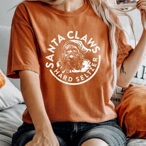 May include: An orange short-sleeved t-shirt with a white circular graphic. The graphic features a drawing of Santa Claus and the words "SANTA CLAUS HARD SELTZER". The shirt is made of a soft material.