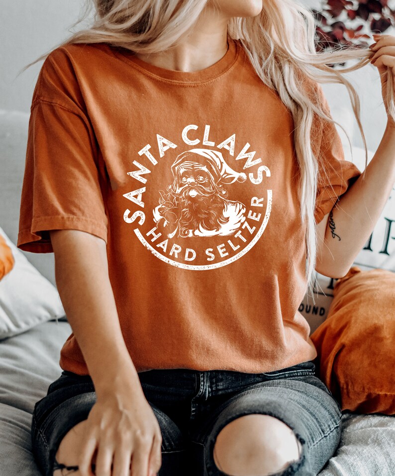 May include: An orange short-sleeved t-shirt with a white circular graphic. The graphic features a drawing of Santa Claus and the words "SANTA CLAUS HARD SELTZER". The shirt is made of a soft material.