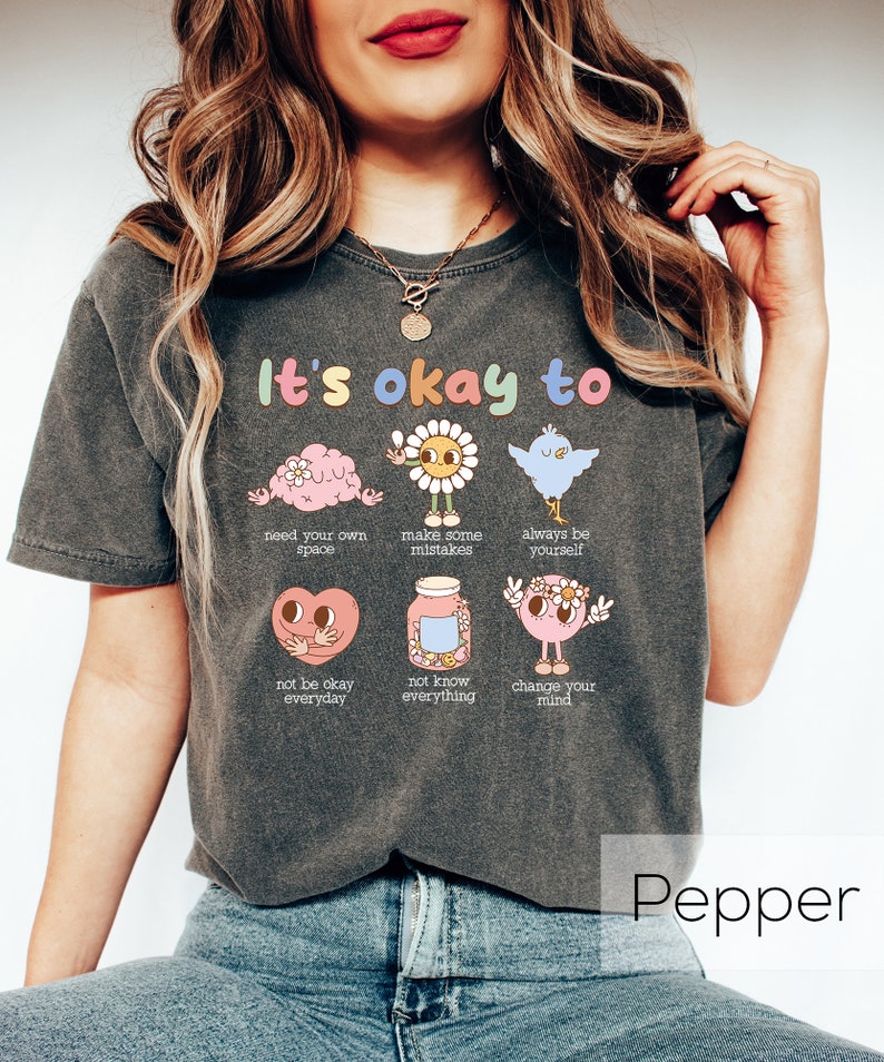 K&ouml;nnte beinhalten: Ein dunkelgraues T-Shirt mit dem Text "It's okay to" und Illustrationen eines Gehirns, einer Blume, eines Vogels, eines Herzens, eines Glases und einer Person. Die Illustrationen sind in Pastellfarben gehalten und haben positive Botschaften.
