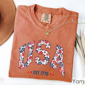 May include: A terracotta-colored t-shirt with the word "USA" in a floral design. The letters are filled with red, white, and blue flowers. Below the word "USA" is the text "EST 1776" with two red stars on each side. A gold necklace is draped across the shirt.