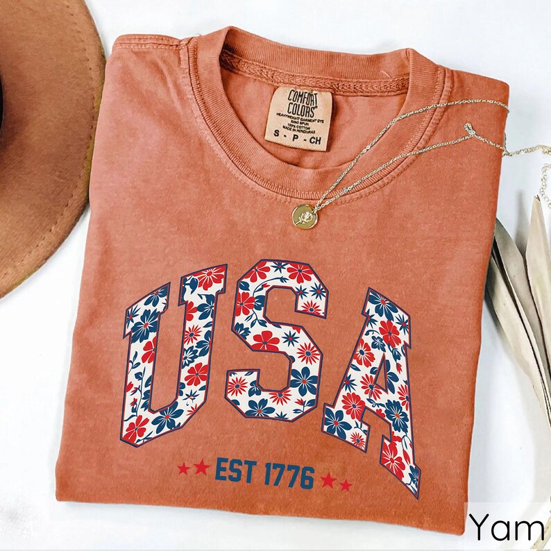 May include: A terracotta-colored t-shirt with the word "USA" in a floral design. The letters are filled with red, white, and blue flowers. Below the word "USA" is the text "EST 1776" with two red stars on each side. A gold necklace is draped across the shirt.