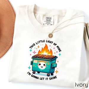 May include: A white t-shirt with a graphic of a green dumpster on fire with the text "This little light of mine, I'm gonna let it shine".