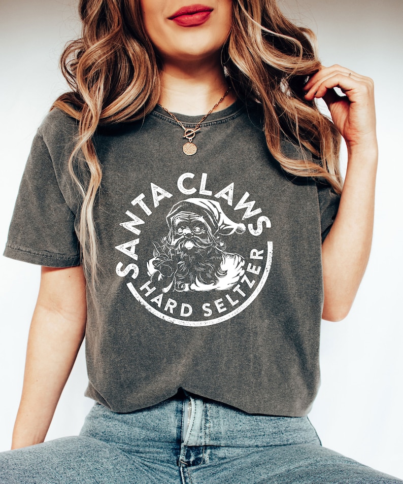 May include: Dark gray t-shirt with a white graphic that reads "SANTA CLAWS HARD SELTZER" in a circular design. The graphic features a cartoon Santa Claus. The shirt is a short-sleeved, crew neck style.