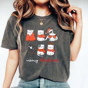 May include: A charcoal gray t-shirt featuring a graphic of six cartoon cats in winter attire, including scarves, a Santa hat, and reindeer antlers. The text below the cats reads meowy christmas in white and red script lettering.