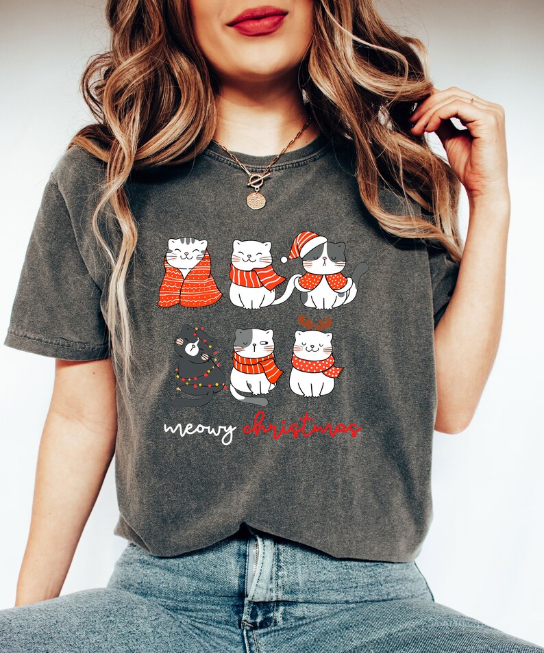 May include: A charcoal gray t-shirt featuring a graphic of six cartoon cats in winter attire, including scarves, a Santa hat, and reindeer antlers. The text below the cats reads meowy christmas in white and red script lettering.