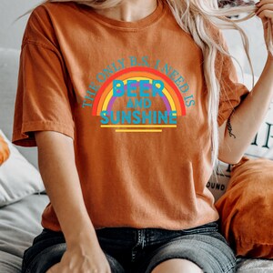 May include: Burnt orange t-shirt with the text "THE ONLY B.S. I NEED IS BEER AND SUNSHINE" in a rainbow design. The shirt is a short-sleeved, casual style.