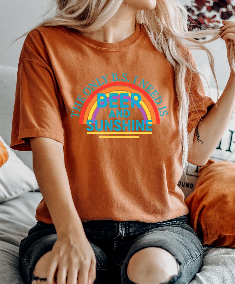 May include: Burnt orange t-shirt with the text "THE ONLY B.S. I NEED IS BEER AND SUNSHINE" in a rainbow design. The shirt is a short-sleeved, casual style.