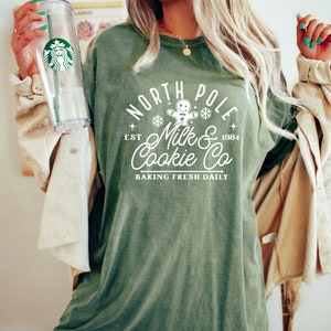 May include: An olive green t-shirt with the text "North Pole Milk & Cookie Co" in white, featuring a gingerbread man and snowflakes. The shirt has a vintage look and feel. A clear Starbucks tumbler with a green straw is held in the left hand.