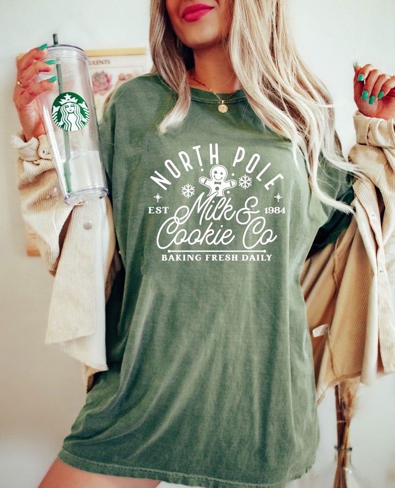 May include: An olive green t-shirt with the text "North Pole Milk & Cookie Co" in white, featuring a gingerbread man and snowflakes. The shirt has a vintage look and feel. A clear Starbucks tumbler with a green straw is held in the left hand.