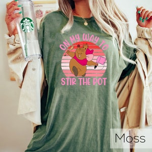 May include: A green t-shirt featuring a vintage-style graphic of a cartoon mouse holding a spoon and a pot. The text on the shirt reads "On my way to stir the pot".