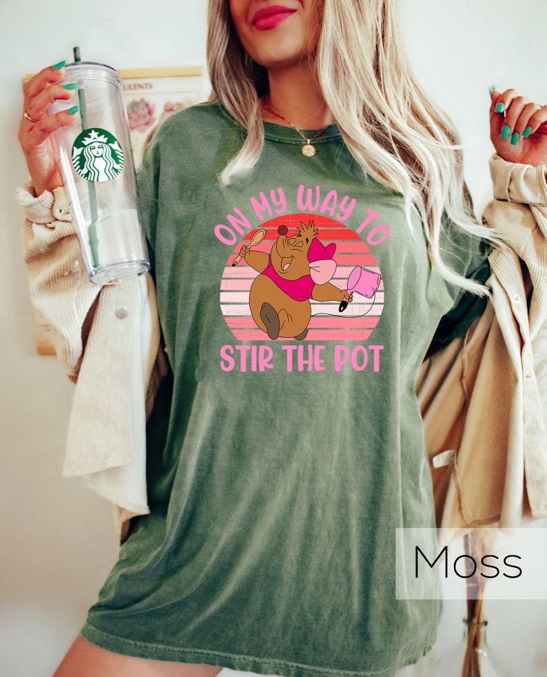 May include: A green t-shirt featuring a vintage-style graphic of a cartoon mouse holding a spoon and a pot. The text on the shirt reads "On my way to stir the pot".