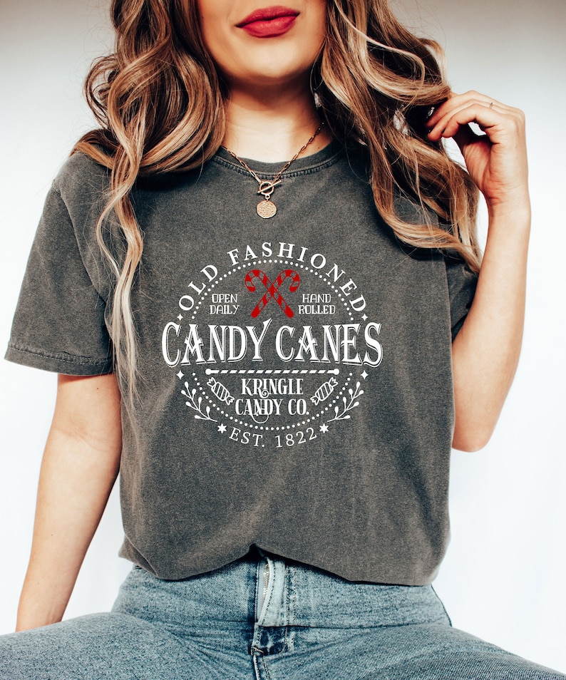 Pu&ograve; includere: T-shirt grigio scuro con grafica circolare che recita "Old Fashioned Candy Canes" con illustrazioni di bastoncini di zucchero rossi. Il testo include anche "Kringle Candy Co. Est. 1822". La maglietta &egrave; di vestibilit&agrave; ampia.