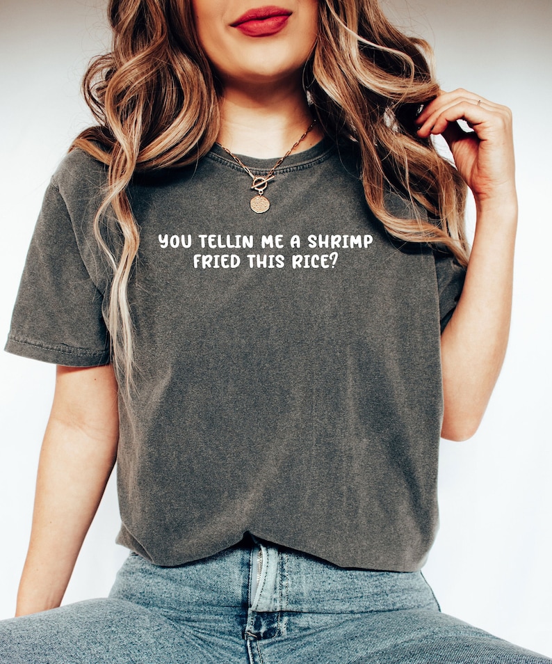 May include: Dark grey t-shirt with the text "YOU TELLIN ME A SHRIMP FRIED THIS RICE?" in white. The shirt is a relaxed fit and is paired with blue jeans. The model is wearing a gold necklace.