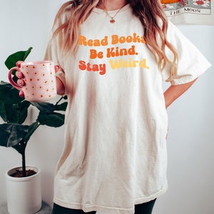 K&ouml;nnte beinhalten: Ein wei&szlig;es Oversize-T-Shirt mit dem Text "Read Books. Be Kind. Stay Weird." in orange, gelber und roter Schrift.