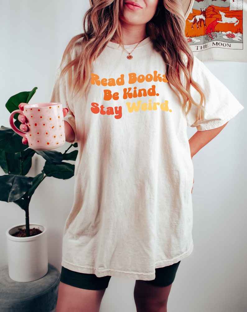 K&ouml;nnte beinhalten: Ein wei&szlig;es Oversize-T-Shirt mit dem Text "Read Books. Be Kind. Stay Weird." in orange, gelber und roter Schrift.