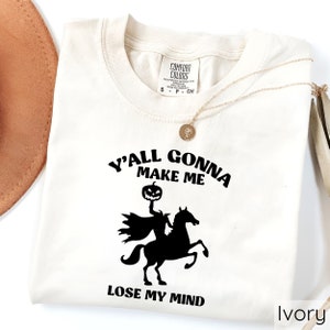 May include: A white t-shirt with a black graphic of a headless horseman riding a horse. The graphic includes the text "Y'all Gonna Make Me Lose My Mind". The shirt is labeled "Ivory".