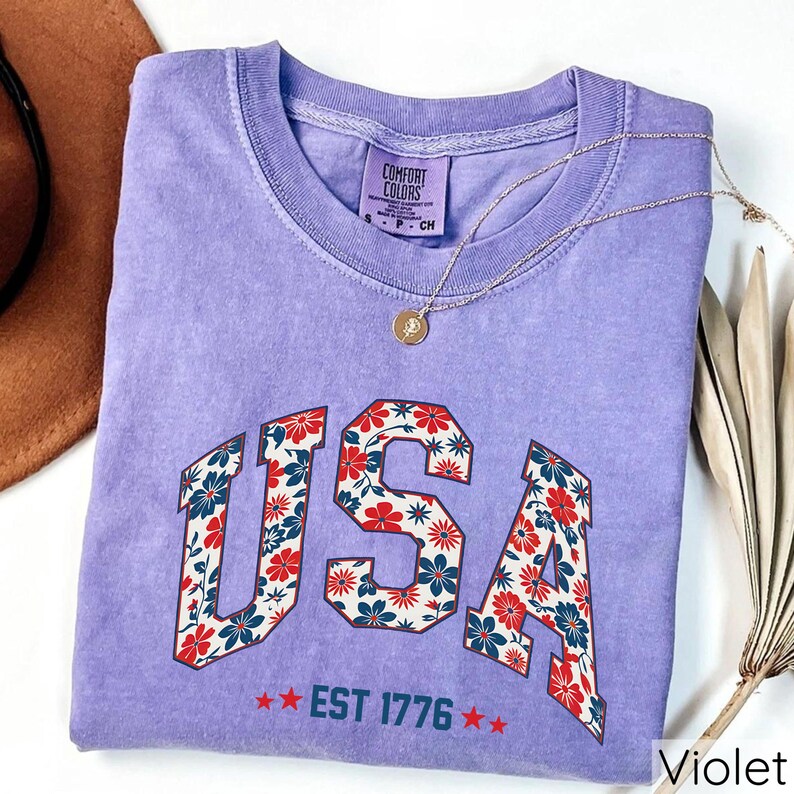 May include: A violet Comfort Colors t-shirt with the word "USA" in a floral design. The shirt also features the text "EST 1776" and is accessorized with a gold necklace. The shirt is a size small.
