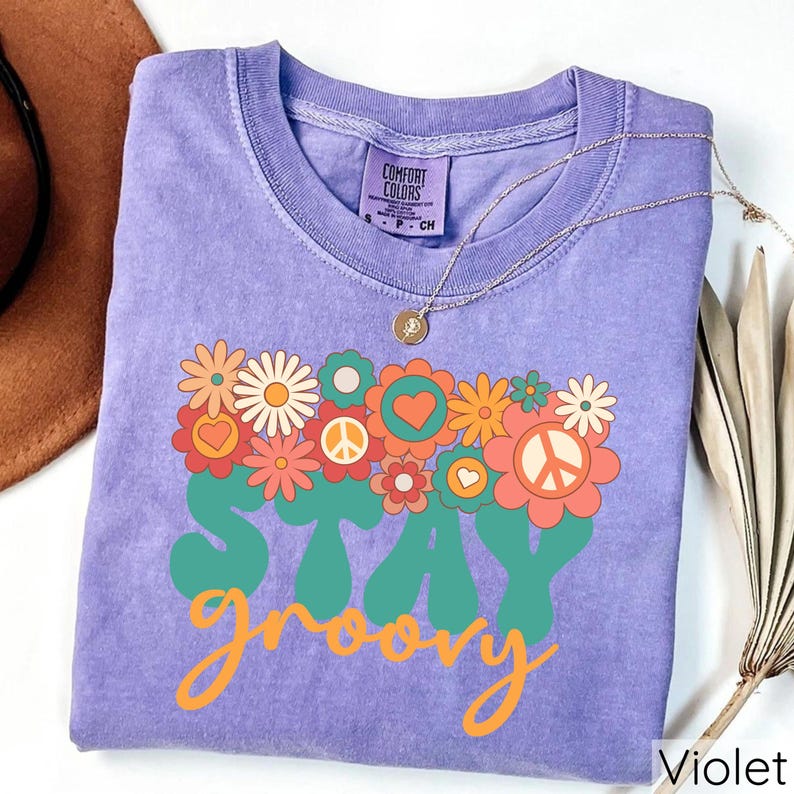 May include: A purple t-shirt with a vintage-style graphic that says "Stay groovy" with a peace sign and flowers. The shirt is made by Comfort Colors.