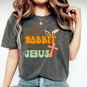 May include: A gray t-shirt with a retro-style graphic that says "Silly Rabbit, Easter is for Jesus". The graphic features a pink bunny with a cross and is in orange, pink, and green.
