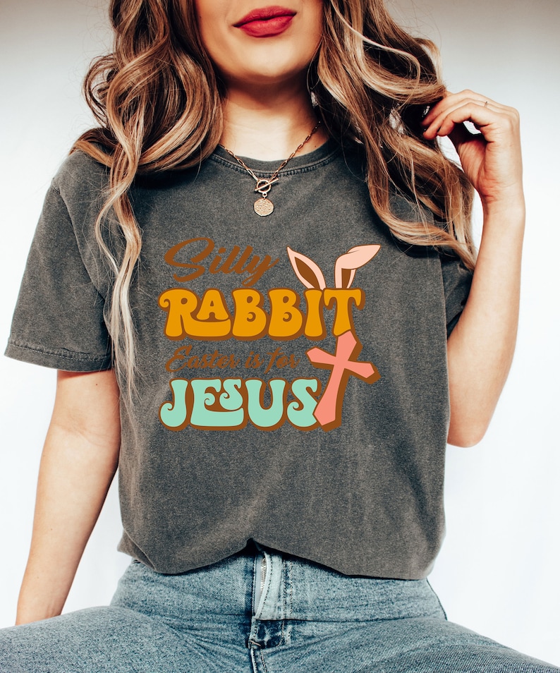 May include: A gray t-shirt with a retro-style graphic that says "Silly Rabbit, Easter is for Jesus". The graphic features a pink bunny with a cross and is in orange, pink, and green.