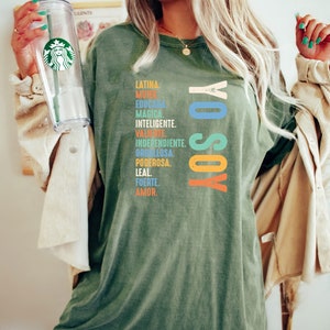 May include: A green t-shirt with the text "YO SOY" in rainbow colors and a list of Spanish words describing a woman's qualities.