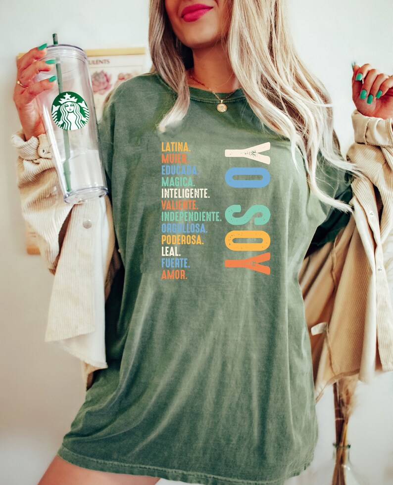 May include: A green t-shirt with the text "YO SOY" in rainbow colors and a list of Spanish words describing a woman's qualities.